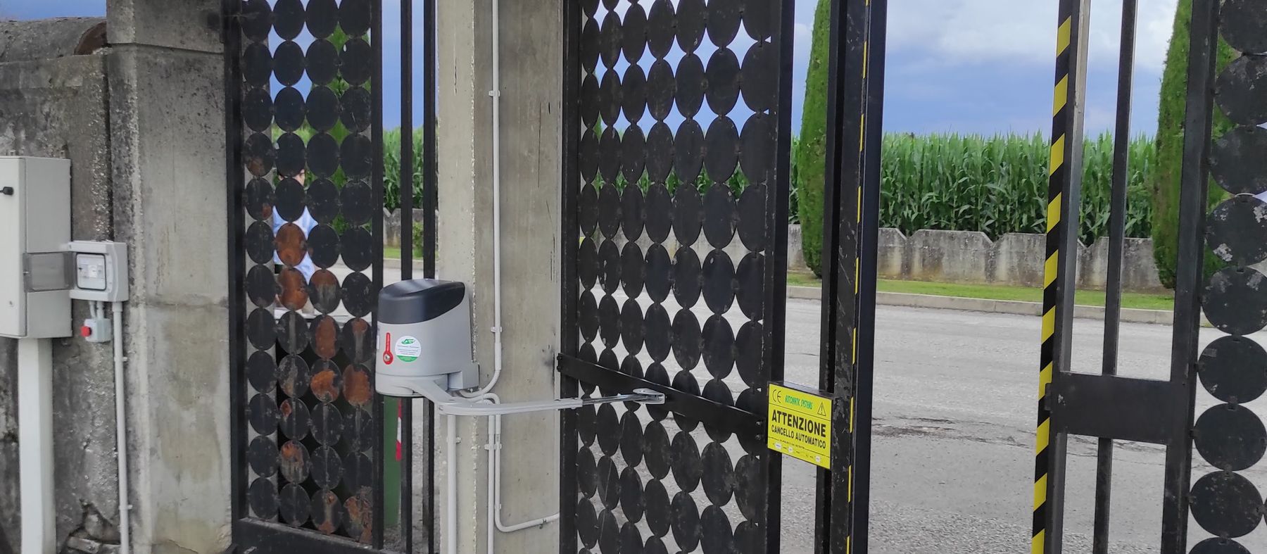 Cancello a due ante motorizzato con automazione Nice, lampeggiante e cartello di avvertimento, installato all'ingresso di un cimitero
